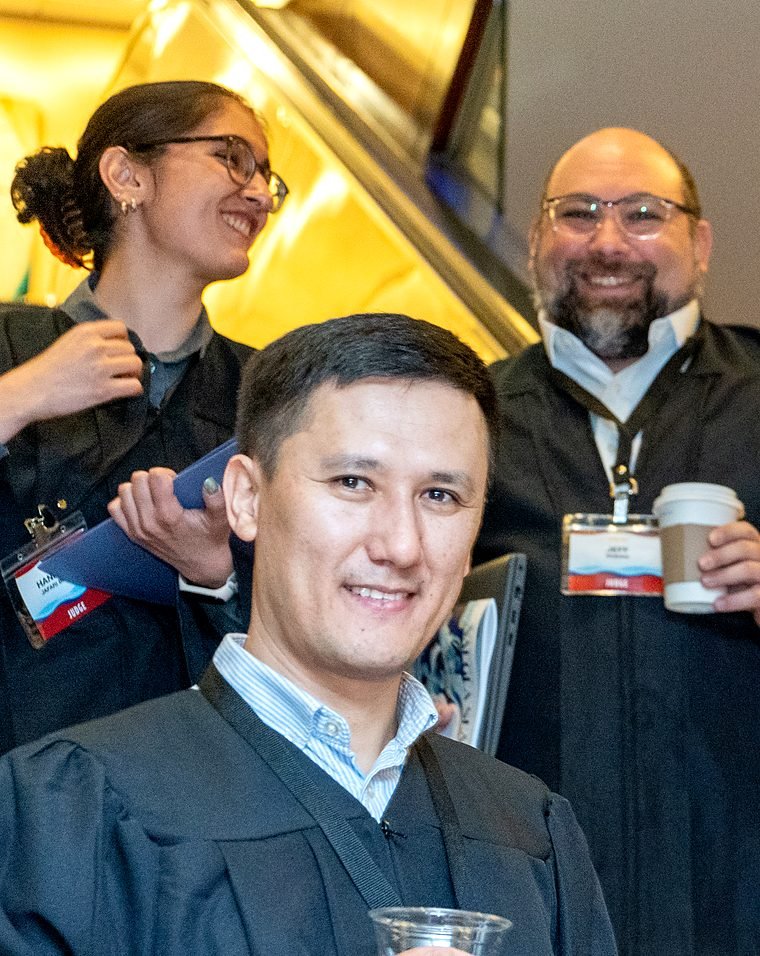 A man in a judge's robe smiles at the camera, holding a plastic cup, with two smiling judges in robes standing behind him.