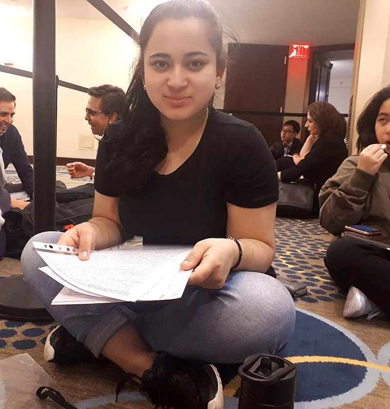 A student smiles at the camera while sitting on the floor with her case papers, other competitors preparing behind her.