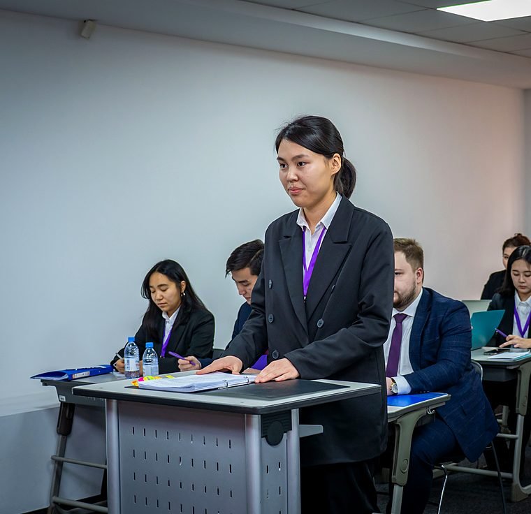 A student stands at her desk to present her argument during an oral round, teammates seated behind her.