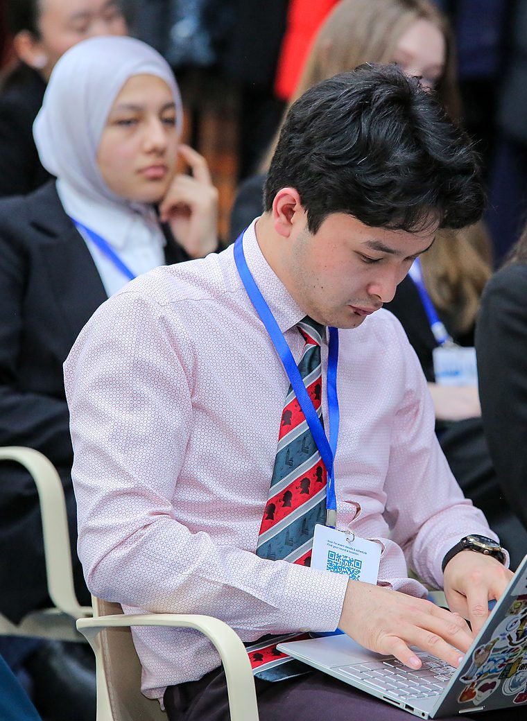 A student in a pink shirt and lanyard types on a laptop, other participants seated around him.