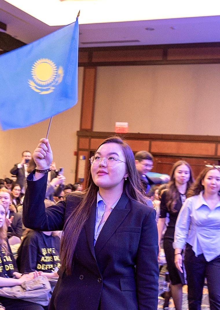 A student holds up the flag of Kazakhstan in a crowded conference hall.