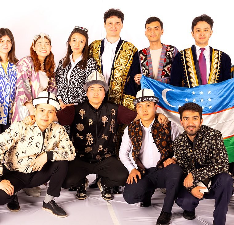 A team in traditional costumes poses together with the flag of Uzbekistan.