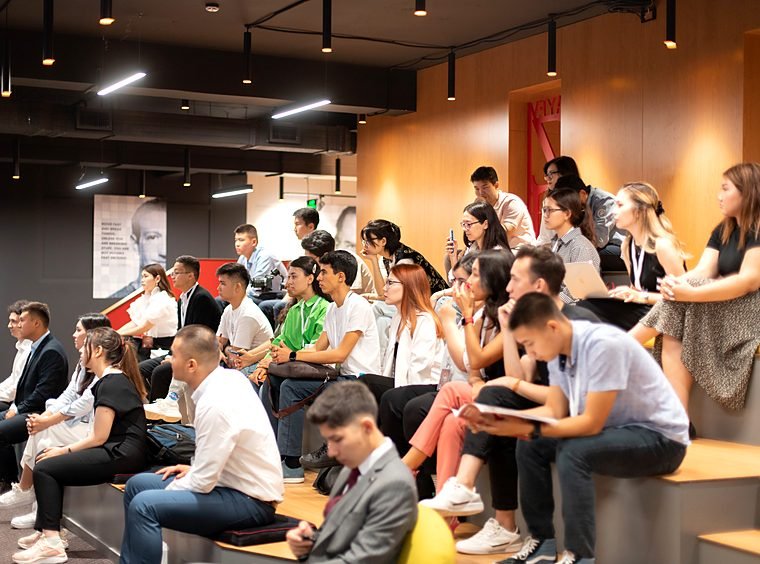 Students seated on tiered steps and chairs listen to a session in a large room.