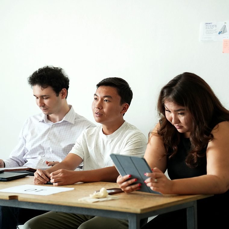 Three students discuss around a table, one of them holding a tablet.