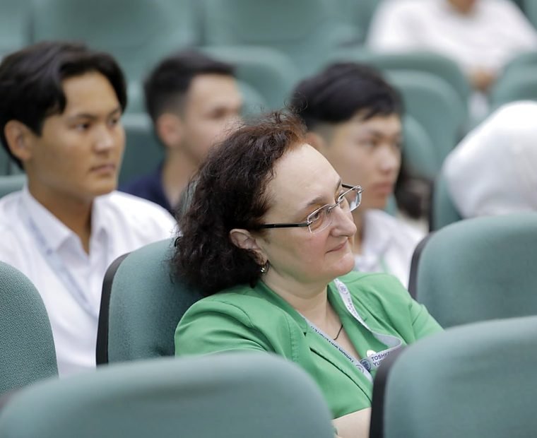 A woman in a green jacket and lanyard sits among students in the audience at the Summer School.
